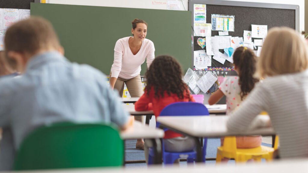Female teacher leaning on desk talking to students, from view of student